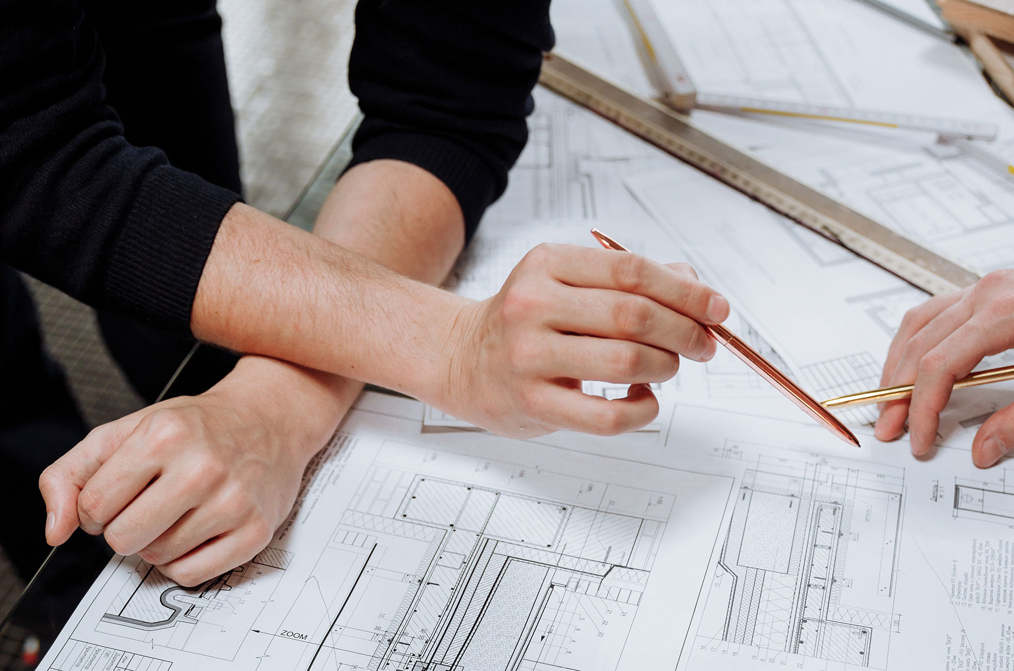 Two hands holding pens hover over detailed architectural drawings, suggesting collaboration or planning on a construction or design project.